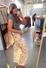 Load image into Gallery viewer, A woman taking a mirror selfie in a clothing boutique, wearing a sage green short-sleeve top and cream wide-leg pants with a small bird print.