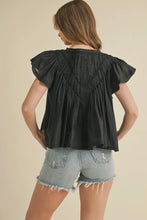Load image into Gallery viewer, Woman wearing a black blouse with ruffled sleeves and denim shorts on a beige background