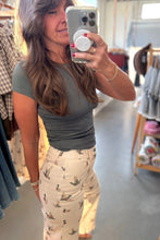 Load image into Gallery viewer, A woman takes a mirror selfie in a clothing store, wearing a dark olive green short-sleeve top and beige pants with a small bird print.