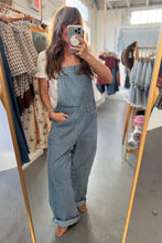 Load image into Gallery viewer, A woman takes a mirror selfie in a clothing boutique wearing a blue and white vertically striped overall jumpsuit. The background shows racks of other clothes and a concrete floor.