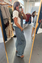 Load image into Gallery viewer, A woman takes a mirror selfie in a clothing boutique, wearing a white ruffled short-sleeve top and blue-and-white vertically striped wide-leg overalls. Clothing racks are visible in the background.