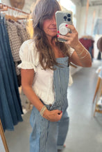 Load image into Gallery viewer, A woman takes a mirror selfie in a clothing store, wearing a white ruffled short-sleeve top and blue-and-white vertically striped overall-style pants. Clothing racks and store fixtures are visible in the background.