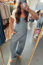 Load image into Gallery viewer, A woman takes a mirror selfie in a clothing boutique wearing blue and white vertically striped denim overall jumpsuit with a white short-sleeve t-shirt underneath.