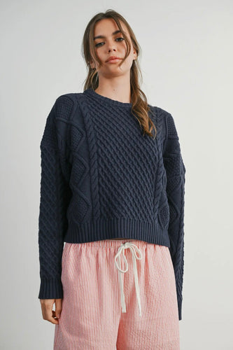 Woman wearing a dark blue cable knit sweater and pink pants with a white background