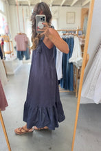 Load image into Gallery viewer, A woman takes a mirror selfie in a clothing boutique wearing a sleeveless, dark navy blue maxi dress with ruffled tiers and tie details at the shoulders, paired with brown strappy sandals.