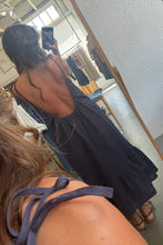 Load image into Gallery viewer, A woman takes a mirror selfie in a clothing store to show off a black backless maxi dress. The photo is taken from a first-person perspective, showing the back of the model wearing the dress, with clothing racks and a textured white wall visible in the background.