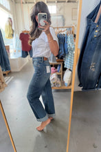 Load image into Gallery viewer, A woman takes a mirror selfie in a clothing boutique wearing a white short-sleeve top and wide-leg blue denim jeans with rolled cuffs. A wooden dress form stands to her right, and other denim garments are hanging in the background.