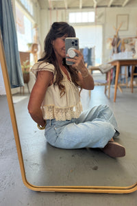 Woman taking a mirror selfie wearing a beige lace top and light blue jeans in a room with wooden furniture.