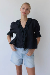 Woman wearing a black ruffled blouse and light blue shorts against a plain background