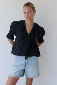Woman wearing a black blouse with ruffles and light blue denim shorts against a white background