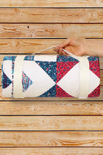 Load image into Gallery viewer, A hand holds a rolled quilt or blanket with a geometric patchwork pattern in red, blue, white, and navy, secured with a cream strap, against a wooden plank background.