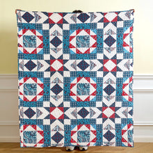 Load image into Gallery viewer, A large quilted blanket or throw with a geometric patchwork pattern in red, white, and blue, held up by a person whose head and feet are mostly hidden behind it, standing in an indoor room with a beige wall and white baseboard.