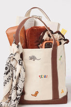Load image into Gallery viewer, A canvas tote bag with brown leather trim and handles, filled with various items including a patterned scarf, a brown wallet, and an orange camera. The bag features small embroidered animal and flower motifs, with the word 'TEXAS' embroidered in colorful letters on the front.