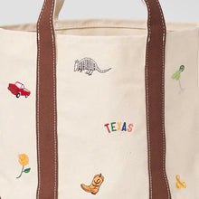Load image into Gallery viewer, A close-up of a canvas tote bag with brown handles and brown side panels, decorated with embroidered Texas-themed motifs including a red car, margarita glass, yellow rose, cowboy boot, armadillo, and the word TEXAS in multicolored letters.