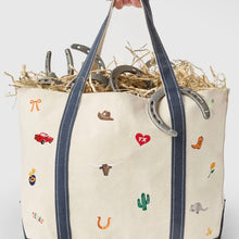 Load image into Gallery viewer, A cream canvas tote bag with navy blue handles and embroidered western-themed motifs, filled with straw and metal horseshoes, held by a hand against a white background.
