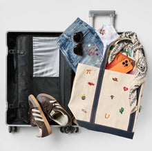 Load image into Gallery viewer, A beige canvas travel organizer tote bag with navy trim and embroidered western-themed motifs is shown packed inside an open silver suitcase, alongside jeans, sunglasses, a white T-shirt, an orange phone case, and brown sneakers on a light gray background.