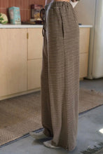 Load image into Gallery viewer, A person wearing loose, wide-leg striped pants in brown and beige, standing indoors near a kitchen counter and rug.