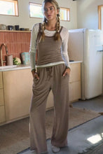 Load image into Gallery viewer, A woman modeling a two-piece lounge set consisting of a brown and beige striped camisole jumpsuit and matching wide-leg pants, styled over a white long-sleeve shirt in a kitchen setting.
