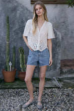 Load image into Gallery viewer, Woman in a white blouse with lace details and denim shorts standing outdoors with cacti in the background.
