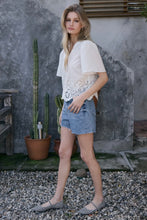 Load image into Gallery viewer, Woman in a white blouse and denim shorts standing outdoors with a cactus plant in the background.