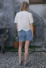 Load image into Gallery viewer, Person wearing a white lace top and denim shorts standing in front of a textured wall with plants.