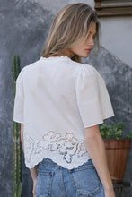 Load image into Gallery viewer, Woman wearing a white blouse with lace details and blue jeans against a gray wall.