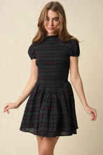 Load image into Gallery viewer, A young woman modeling a short puff-sleeve plaid dress in a dark green, black, and red tartan pattern against a solid beige studio background.