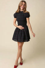 Load image into Gallery viewer, A woman modeling a short black plaid dress with puff sleeves and a flared skirt, paired with red pointed-toe heels, against a plain beige studio background.