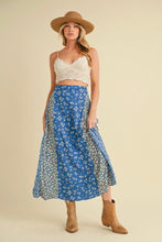 Load image into Gallery viewer, Woman wearing a blue floral skirt, white top, and brown hat on a beige background