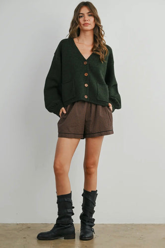Woman wearing a green cardigan, brown shorts, and black boots against a plain background