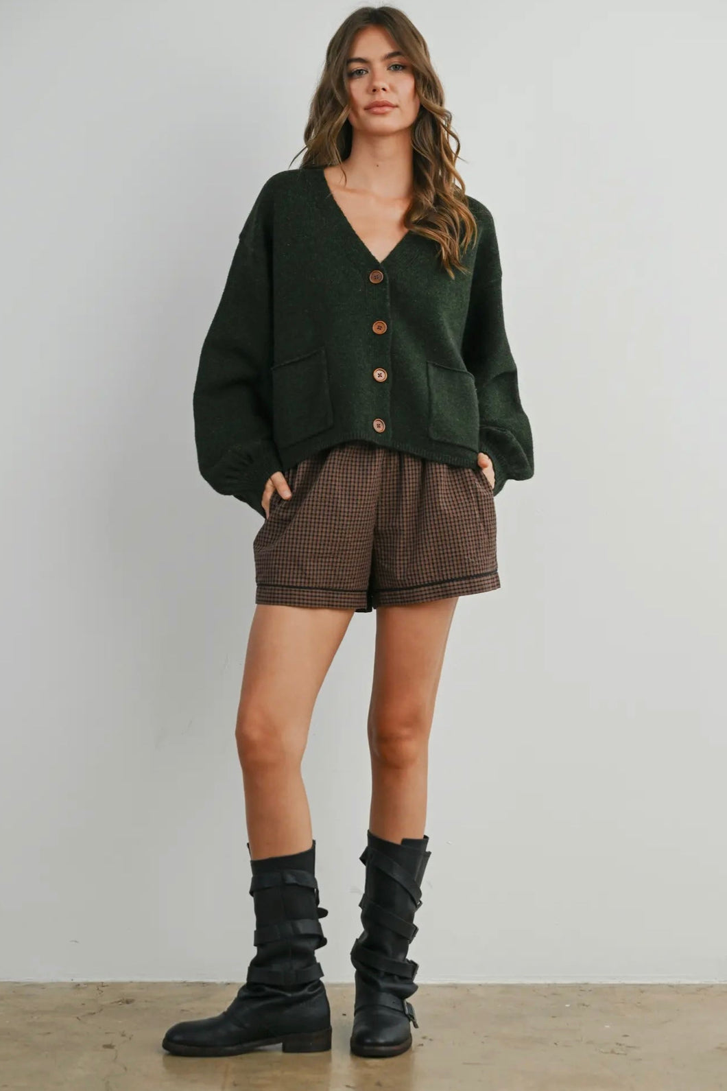Woman wearing a green cardigan, brown shorts, and black boots against a plain background