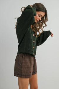Woman wearing a green cardigan and brown checkered shorts on a plain background