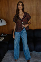 Load image into Gallery viewer, Woman wearing a patterned top and blue jeans standing in a room with wooden walls and a black couch.