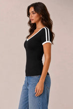 Load image into Gallery viewer, A woman wearing a black short-sleeve V-neck top with white contrast trim and white stripes on the sleeves, paired with blue jeans, photographed against a plain light beige studio background.