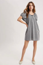 Load image into Gallery viewer, A woman modeling a short gray and white gingham dress with puff sleeves and a gathered neckline, paired with light beige flats, against a plain light gray studio background.