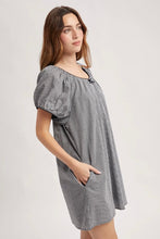 Load image into Gallery viewer, A woman modeling a short-sleeve gray and white striped dress with puff sleeves, a scoop neckline, and side pockets, photographed against a plain light gray studio background.