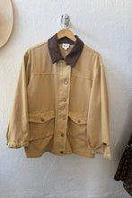 Load image into Gallery viewer, A tan utility jacket with a dark brown collar, hanging on a wooden hanger against a textured white wall.