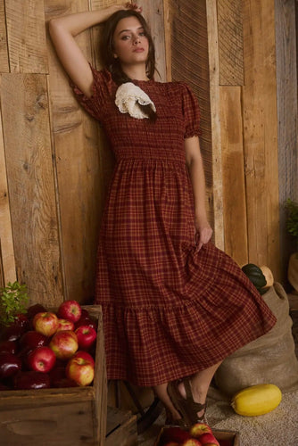 Woman in a red plaid dress standing next to a wooden crate of apples against a rustic wooden wall.