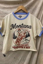 Load image into Gallery viewer, Beige t-shirt with 'Montana Big Sky Country' graphic on a hanger against a textured beige background
