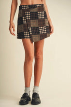 Load image into Gallery viewer, A model wearing a patchwork plaid mini skirt in brown, beige, and black tones, paired with light gray socks and black loafers, against a solid cream background.