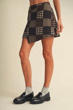 Load image into Gallery viewer, A model wearing a patchwork mini skirt with plaid and checkered patterns in dark brown, beige, and black, paired with white socks and black leather loafers, against a solid light beige background.