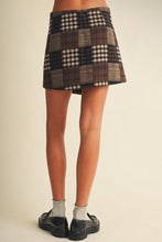 Load image into Gallery viewer, A person shown from the waist down wearing a patchwork plaid mini skirt in brown, beige, and black tones, paired with white socks and black loafers, against a plain beige studio background.