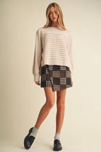 Load image into Gallery viewer, A woman modeling a cream ribbed knit sweater and a brown, black, and beige plaid mini skirt, paired with gray socks and black loafers, against a solid beige studio background.