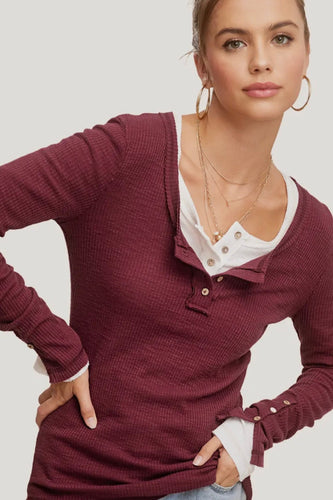Woman wearing a burgundy long-sleeve top with a white shirt underneath on a plain background