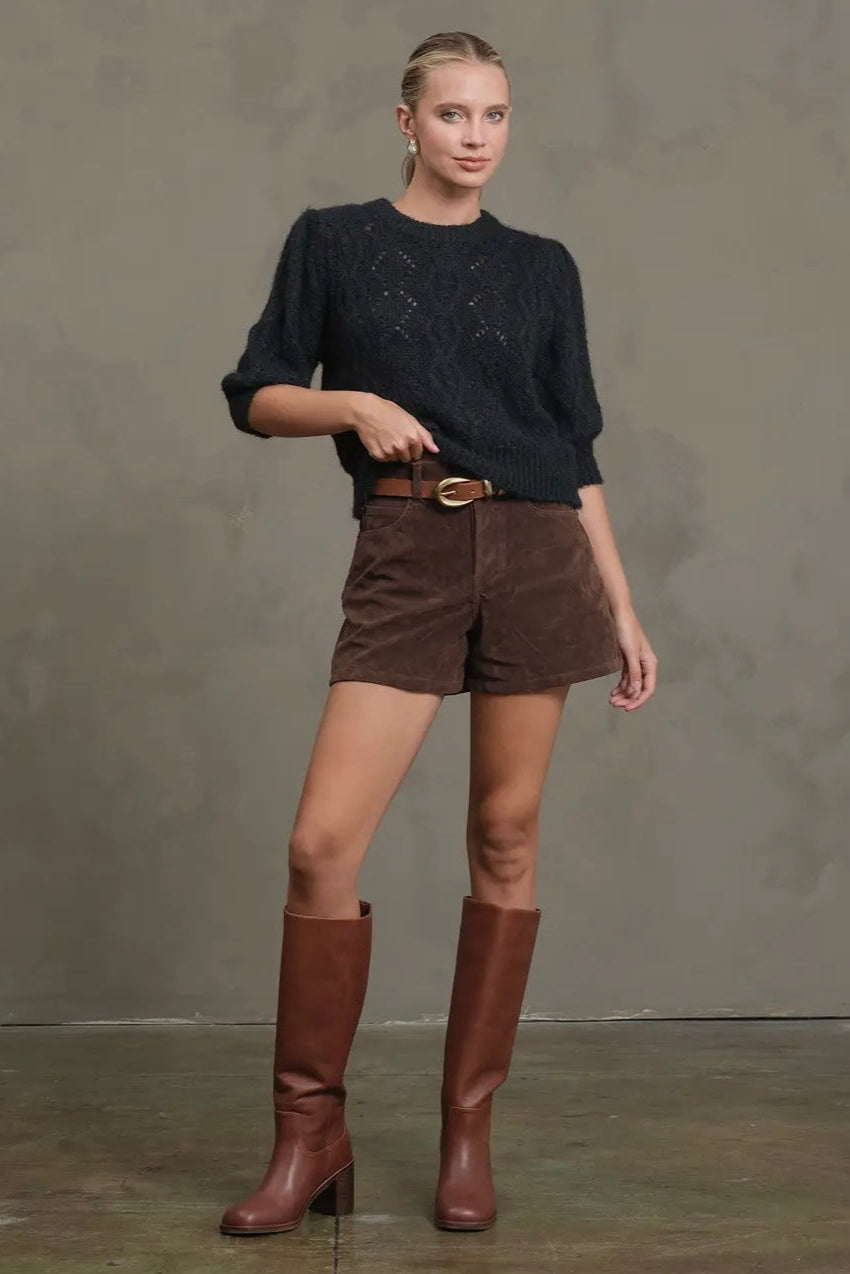 Woman wearing a black sweater, brown shorts, and brown boots against a gray background