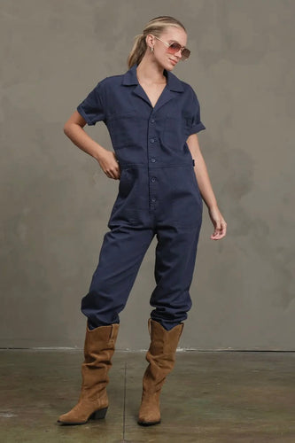 Woman wearing a navy blue jumpsuit and brown boots against a gray background