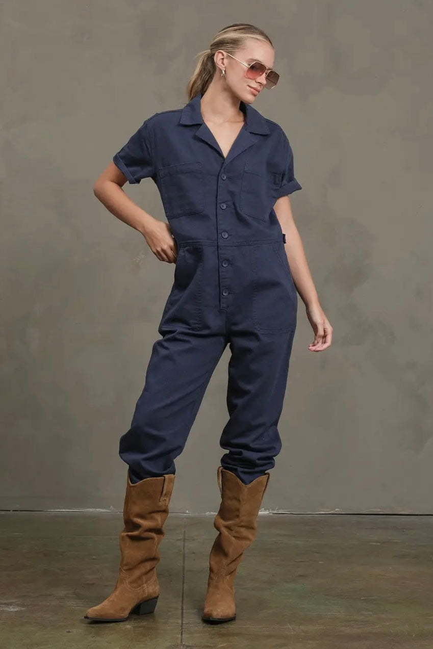 Woman wearing a navy blue jumpsuit and brown boots against a gray background