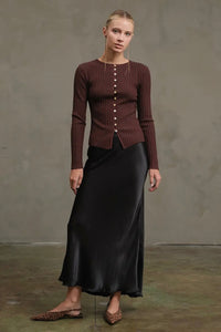 Woman wearing a brown ribbed sweater and black satin skirt against a plain background