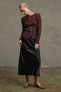 Woman wearing a brown ribbed sweater and black satin skirt against a plain background