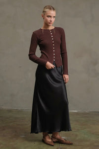 Woman wearing a brown ribbed sweater and black satin skirt against a gray background